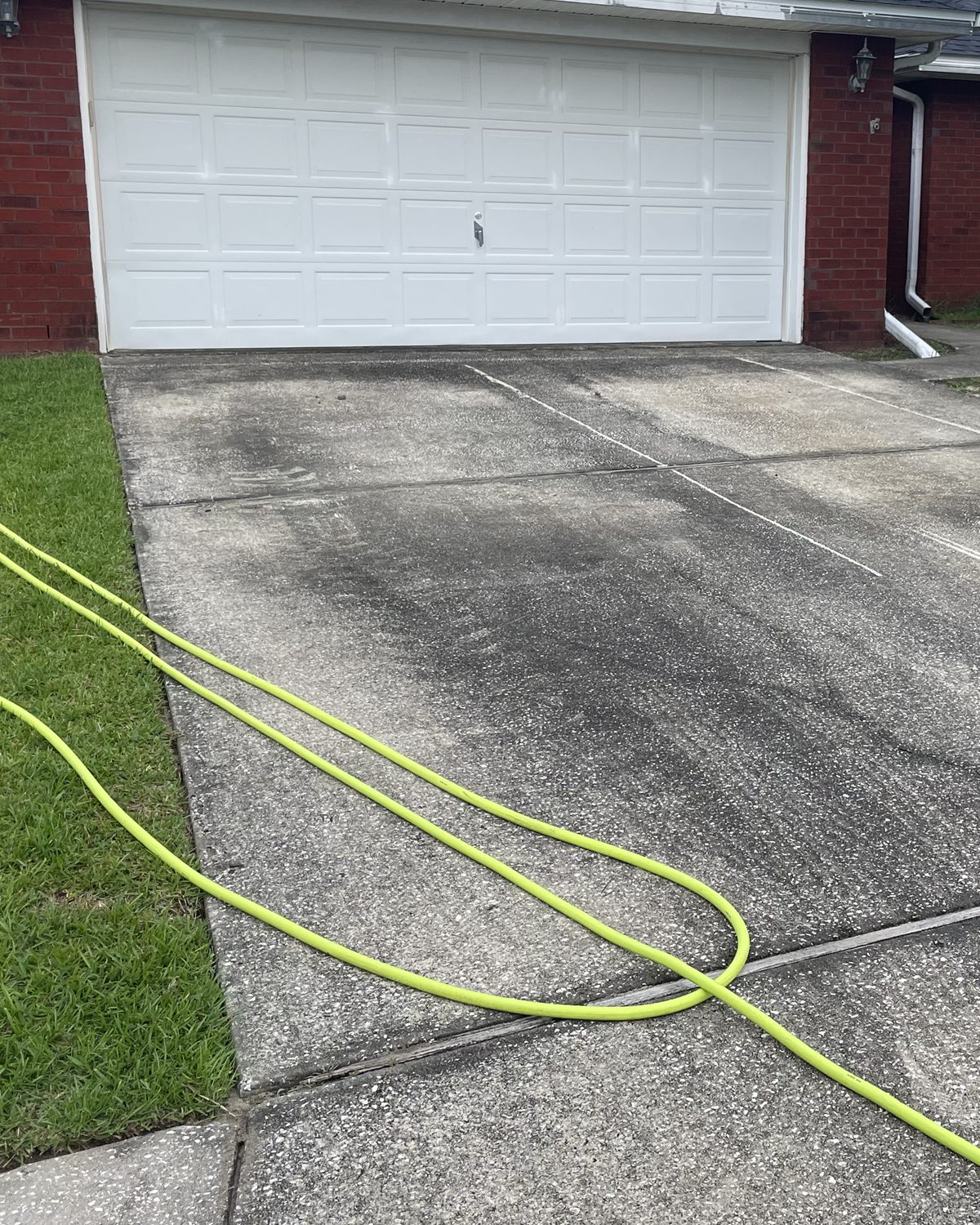 A concrete driveway in front of a red brick garage with a white garage door, dark and stained, with a garden hose laid across it, before cleaning