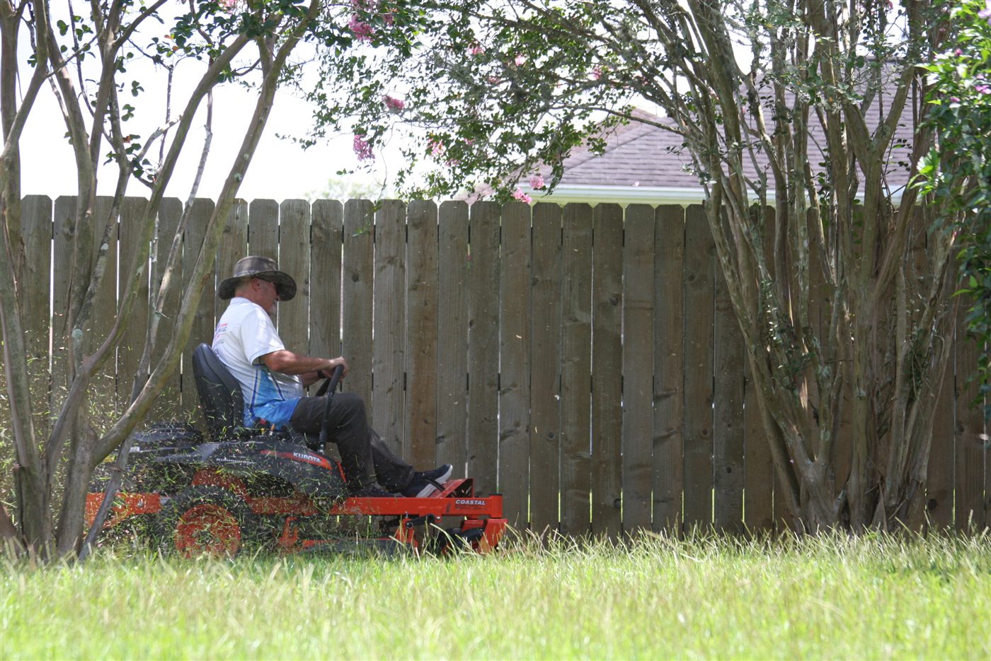 A crew member mowing a lawn along a wooden fence on a zero-turn mower