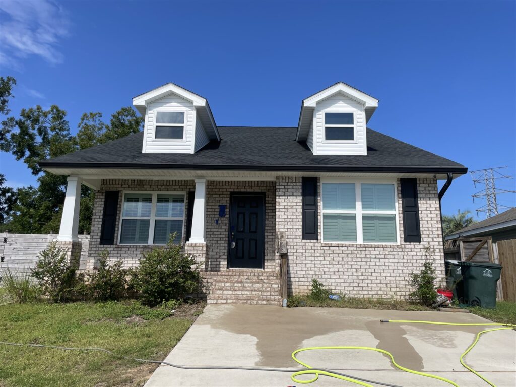 A two-story house with dormers, brick front and a driveway that has been washed, with a hose still laid across it